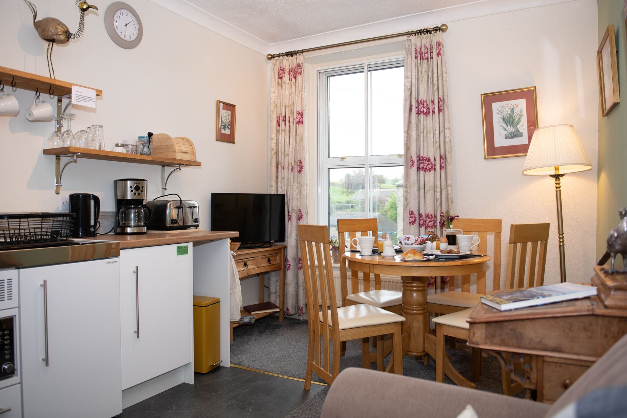 A compact kitchen with a coffee maker, toaster, and a TV. There is a dining area with a table and chairs, and a window with curtains.