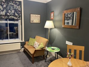 Een gezellige woonkamer met een bruine bank, een klein rond tafeltje, een lamp en een stoel.