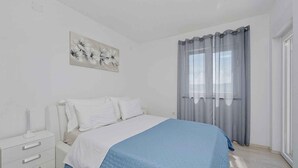 Une chambre à coucher comprenant un lit, une table de chevet, une œuvre d’art accrochée au mur et une fenêtre avec des rideaux.