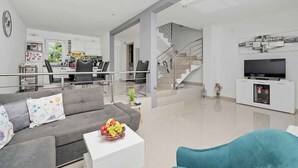 Un salon moderne avec un canapé gris, une table basse blanche ornée de fruits, un fauteuil bleu, un escalier et un téléviseur posé sur un meuble blanc.