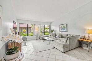 Un salon spacieux avec un canapé gris, une table basse et un lampadaire. De grandes fenêtres offrent une vue sur l’extérieur.