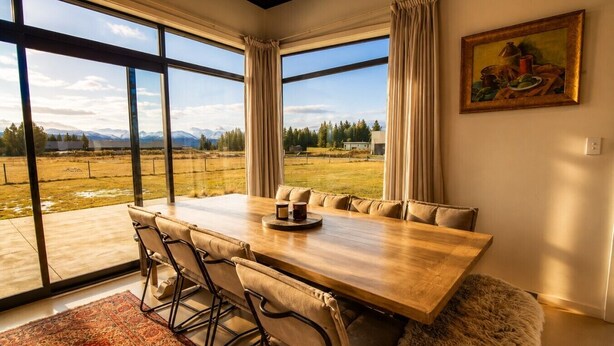 A dining area with a large table and chairs, large windows with curtains, and a painting on the wall.