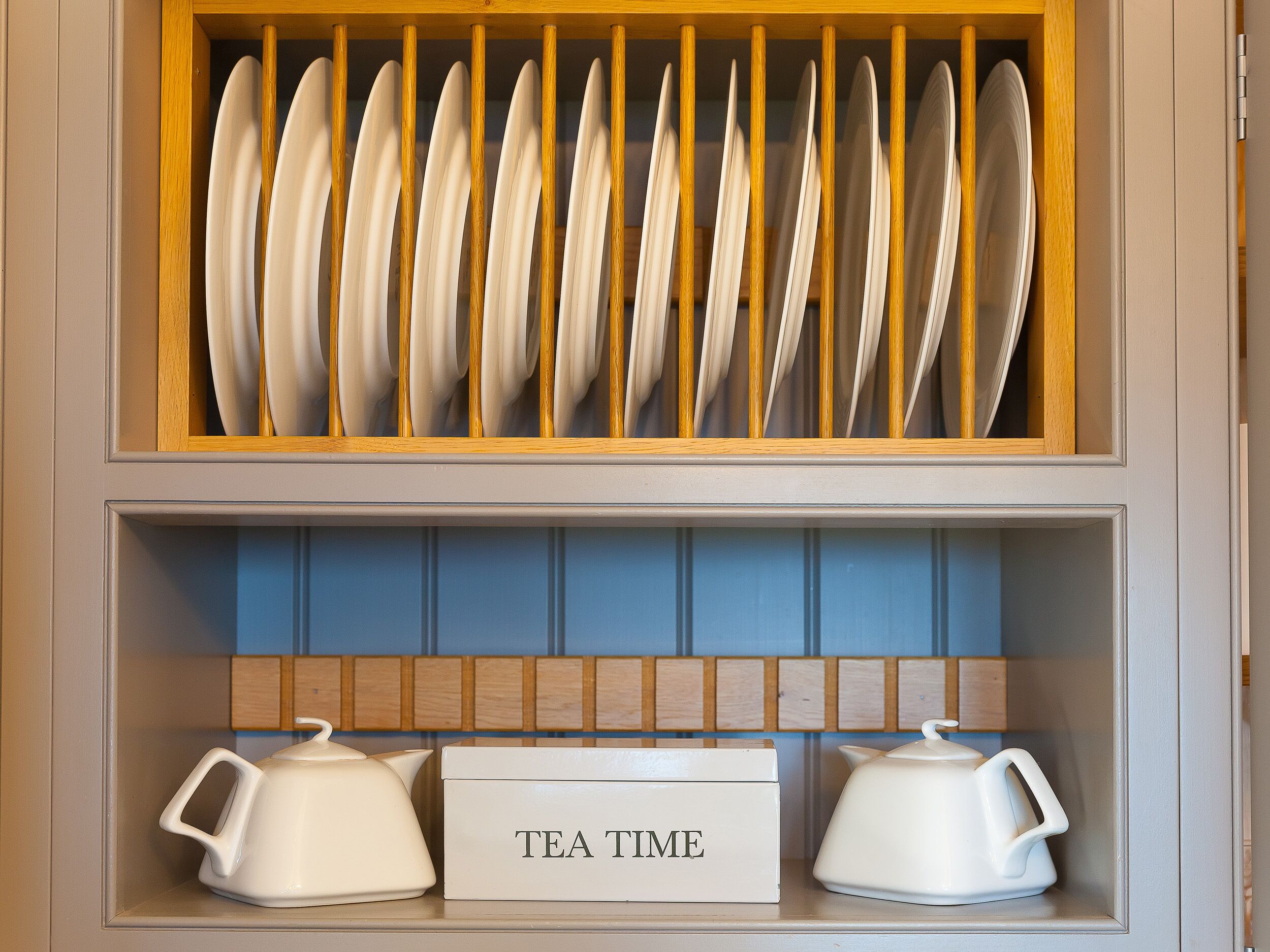 A kitchen with a wooden utensil rack holding white plates and a blue shelf with two white teapots and a sign that reads "TEA TIME".