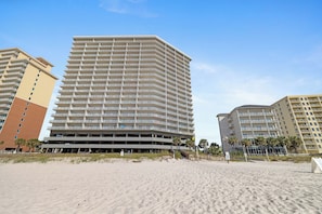 A multi-story hotel with a flat roof and balconies on each floor, located on a sandy beach with palm trees.