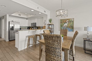 A spacious kitchen with white cabinets, a central island, and a dining area with wicker chairs and a wooden table. - Seawind 1408 by Liquid Life (Gulf Shores)