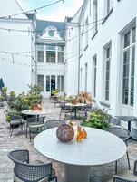 Un patio con mobiliario exterior, incluidas mesas y sillas redondas, rodeado de plantas en macetas y un edificio blanco con ventanas grandes.