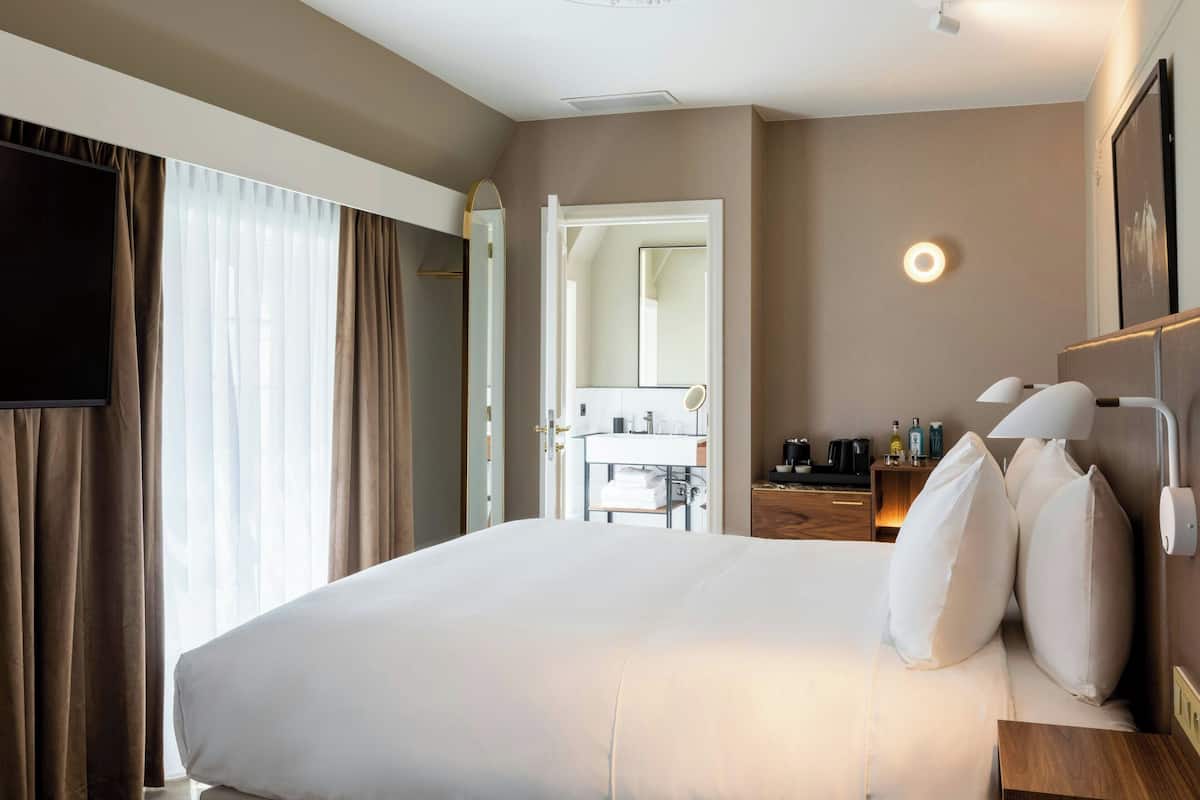 Ein Hotelzimmer mit einem großen Bett, einem Nachttisch mit Lampen, einem Schreibtisch mit Kaffeemaschine und einem durch eine geöffnete Tür einsehbaren Badezimmer.