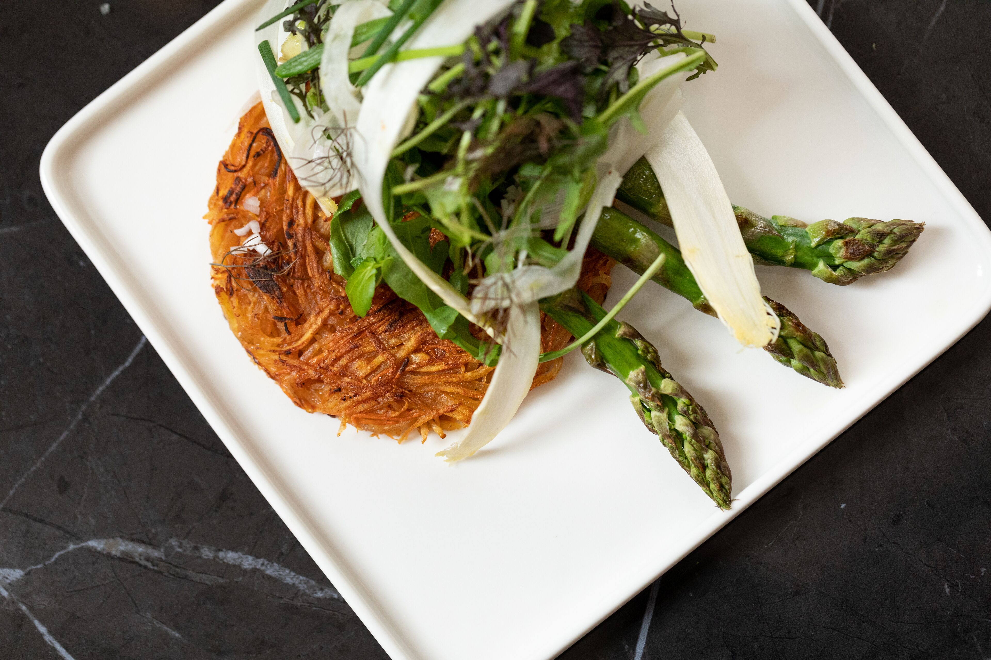 Un plato blanco con un guiso de tortitas de patata crujientes, espárragos y una guarnición de verduras de hoja.
