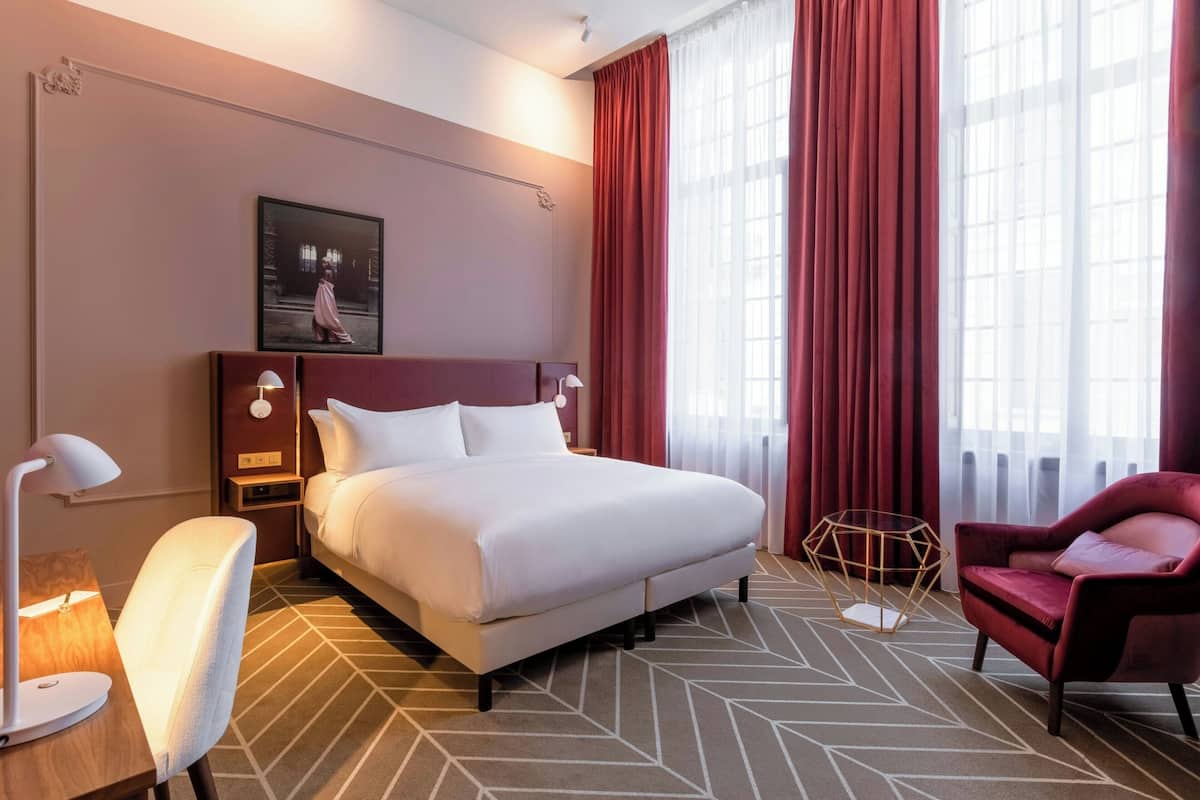 Ein Hotelzimmer mit einem großen Bett, einem Schreibtisch mit Lampe, einem Sessel und einem Fenster mit roten Vorhängen.