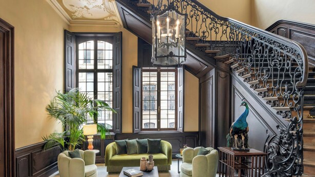 A grand lobby with a staircase, a green sofa, a coffee table, and a peacock statue. - Sapphire House Antwerp, Autograph Collection (Antwerp)