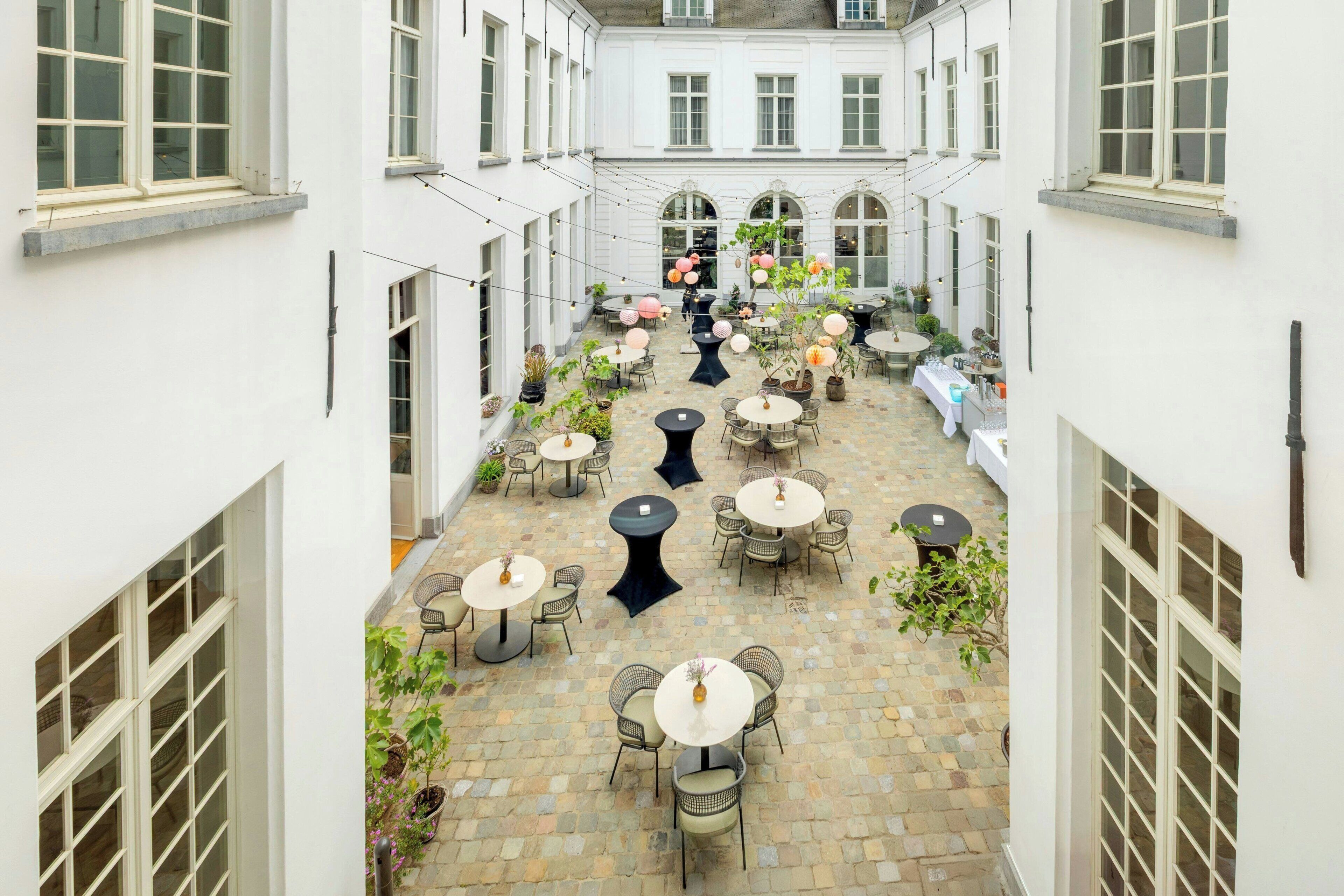 A courtyard with multiple round tables and chairs, surrounded by white walls and large windows.