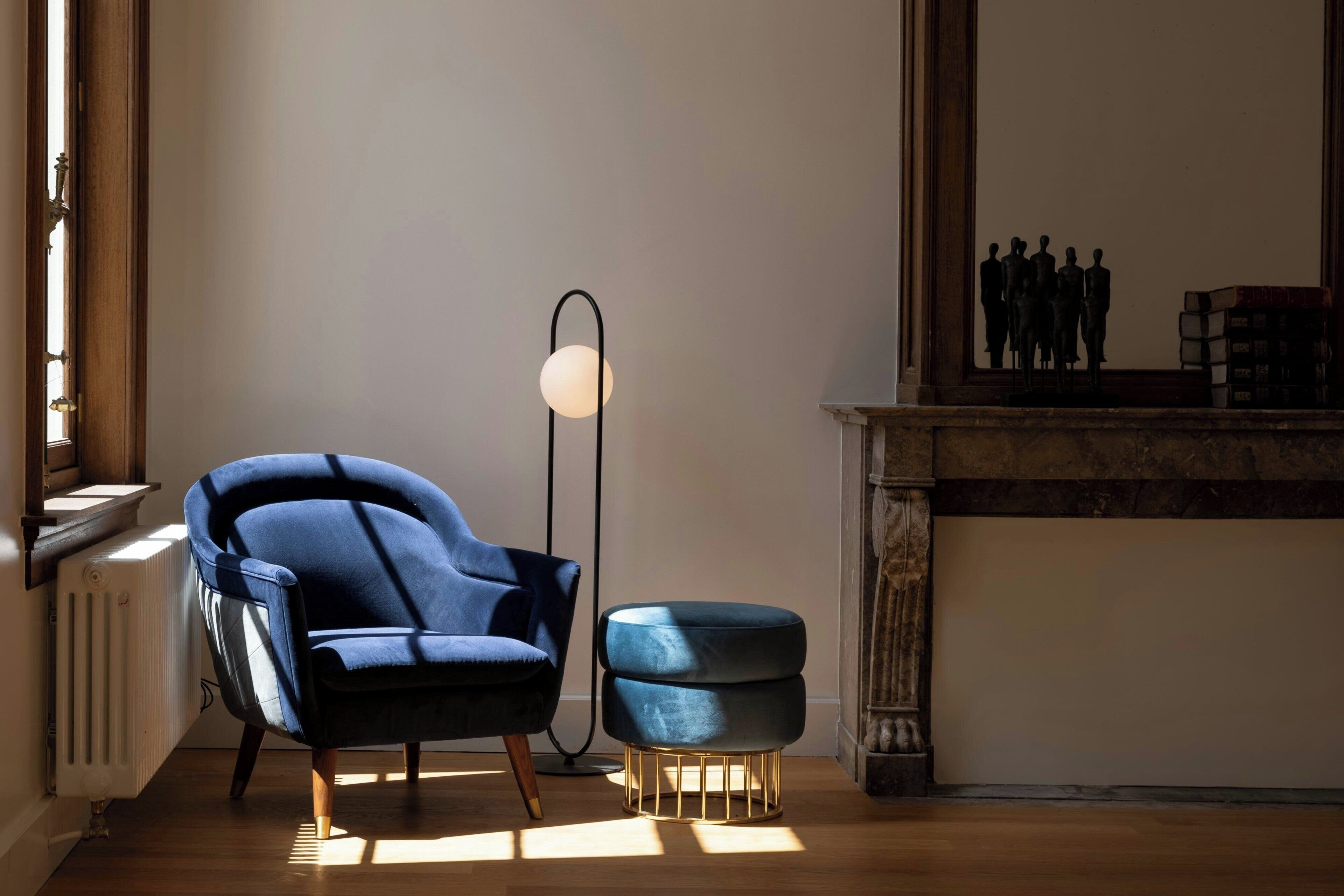 Ein Zimmer mit blauem Sessel, rundem Hocker, Stehlampe und Kamin.