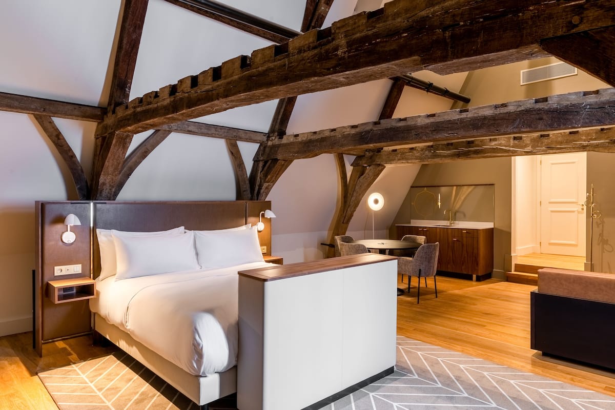 A bedroom with a bed, bedside table, and a desk. There is a wooden ceiling structure and a window.
