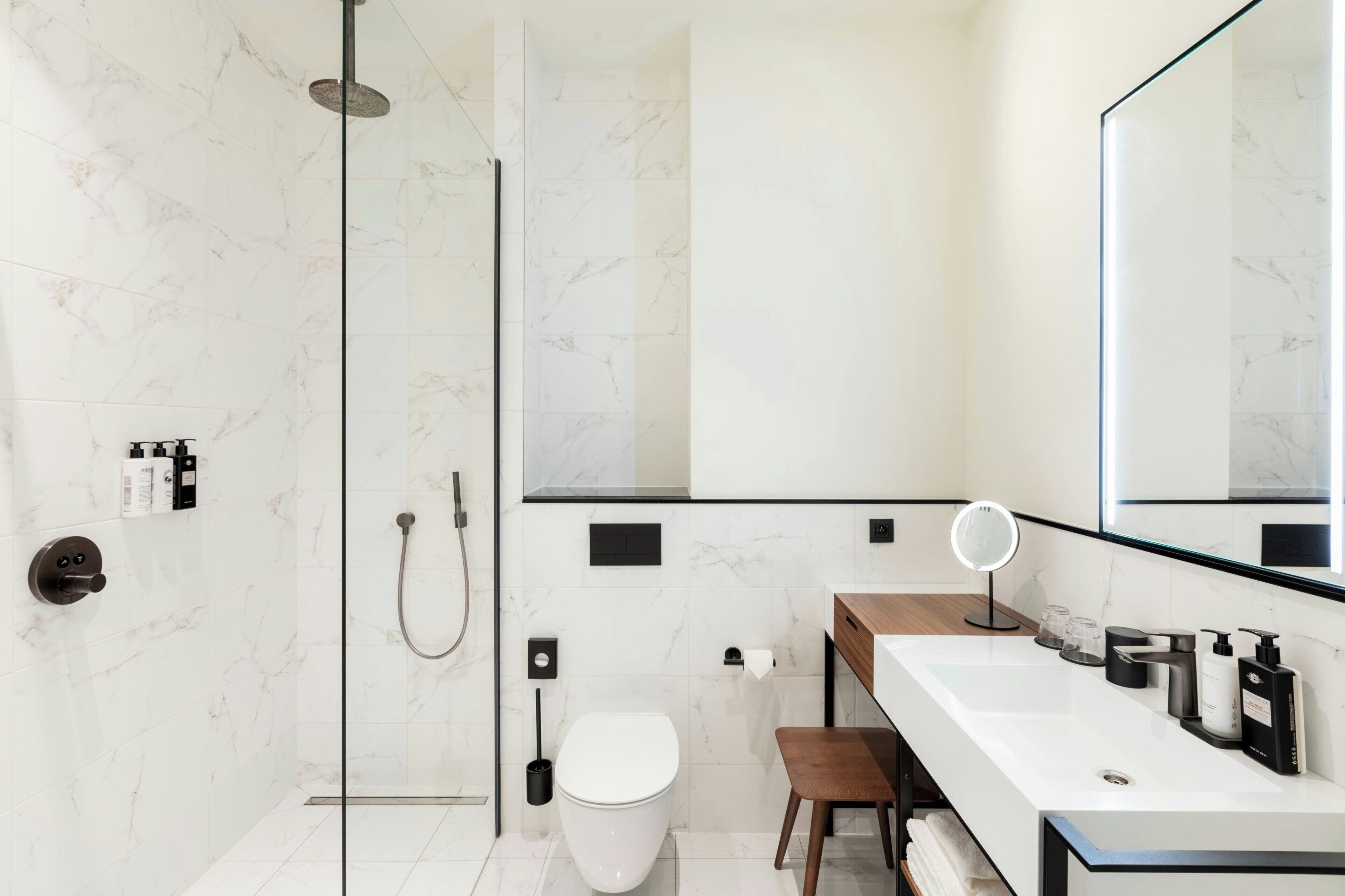 Un baño moderno con cabina de ducha de vidrio, inodoro blanco, tocador de madera con espejo y lavamanos blanco.