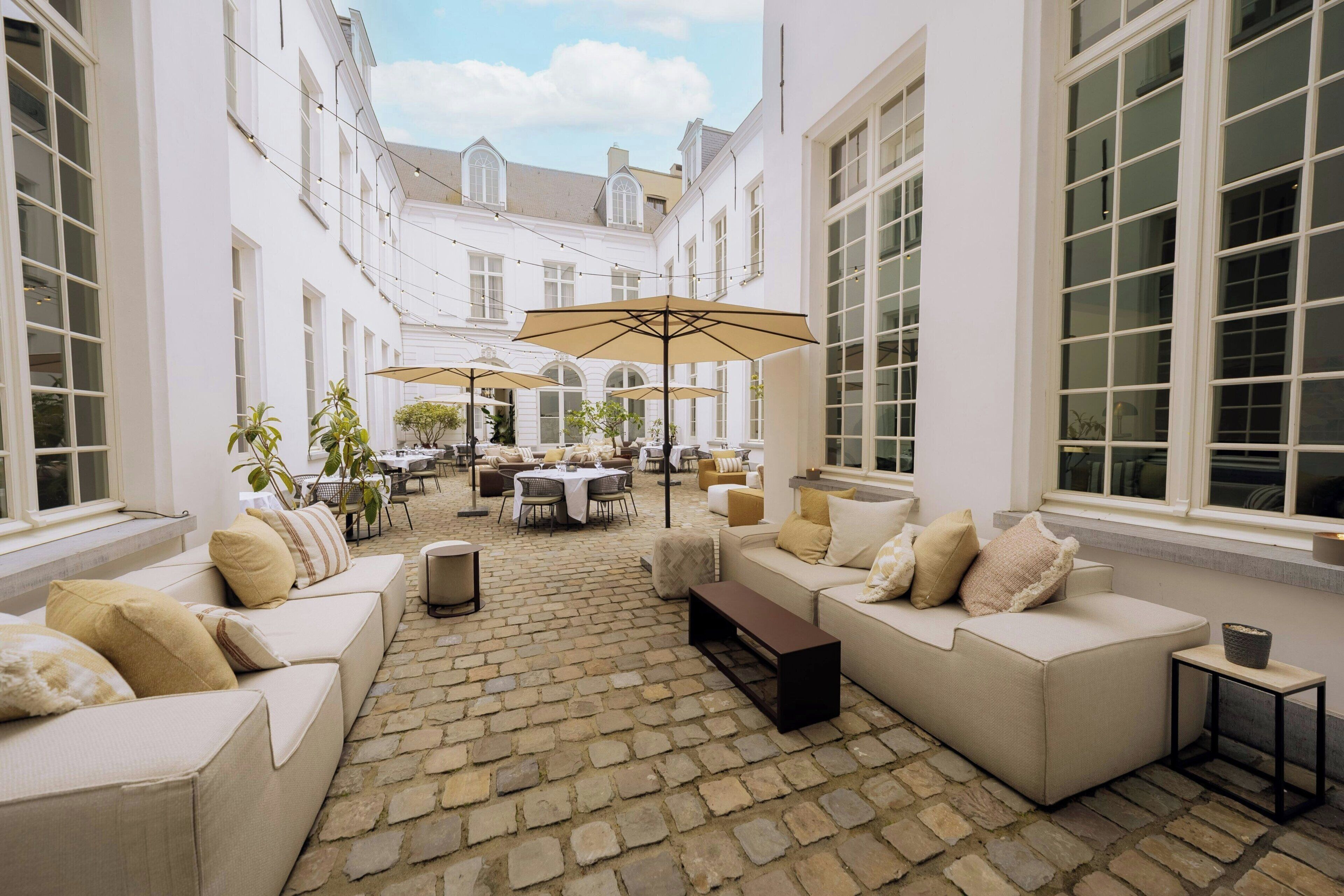 A courtyard with stone flooring, white sofas, outdoor tables, and umbrellas.