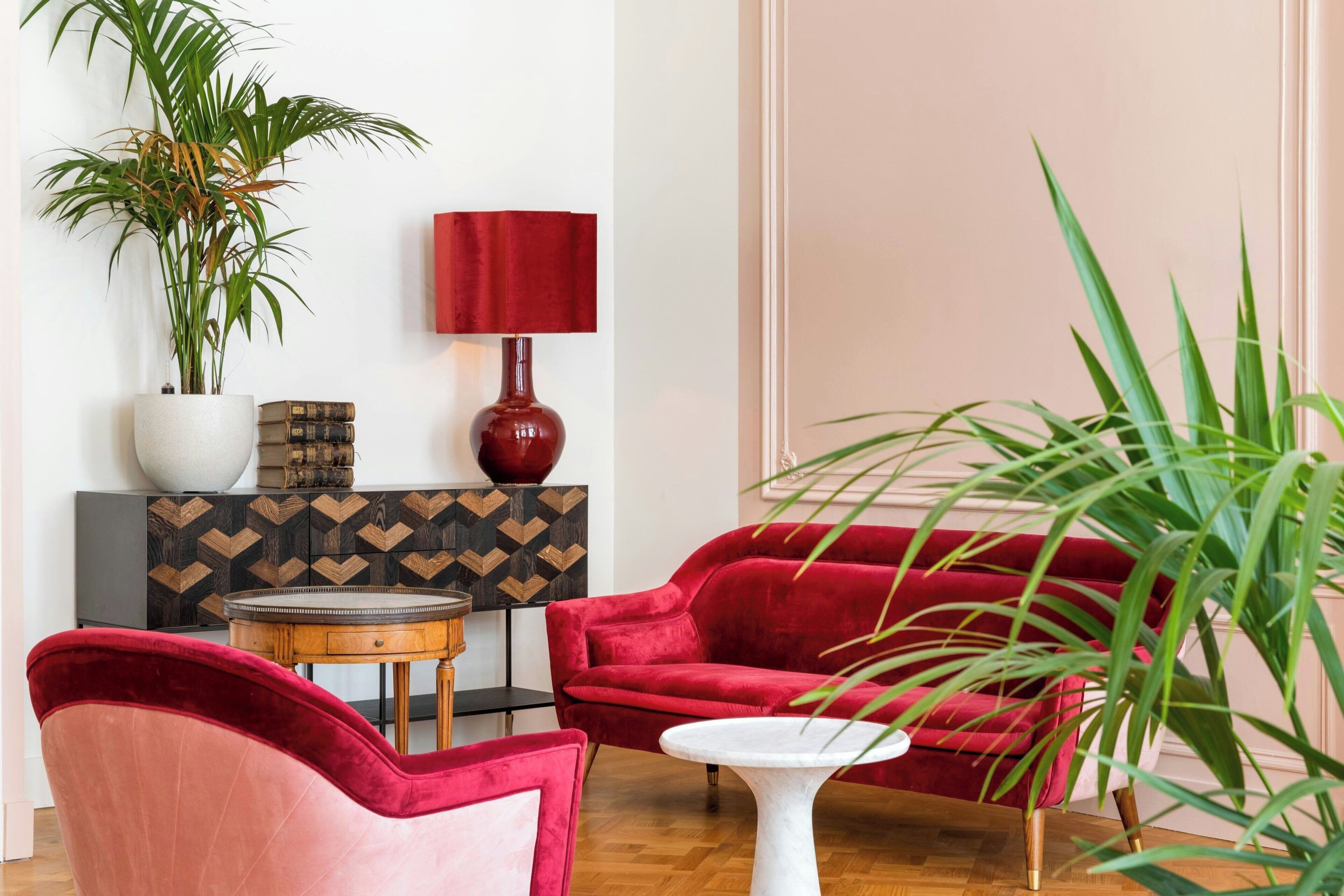 Un canapé en velours rouge, un fauteuil rose, une table blanche et une lampe rouge dans une pièce dont le sol est en bois et qui est agrémentée d’une commode à motifs géométriques.