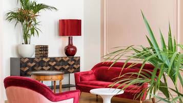 Un canapé en velours rouge, un fauteuil rose, une table blanche et une lampe rouge dans une pièce dont le sol est en bois et qui est agrémentée d’une commode à motifs géométriques.