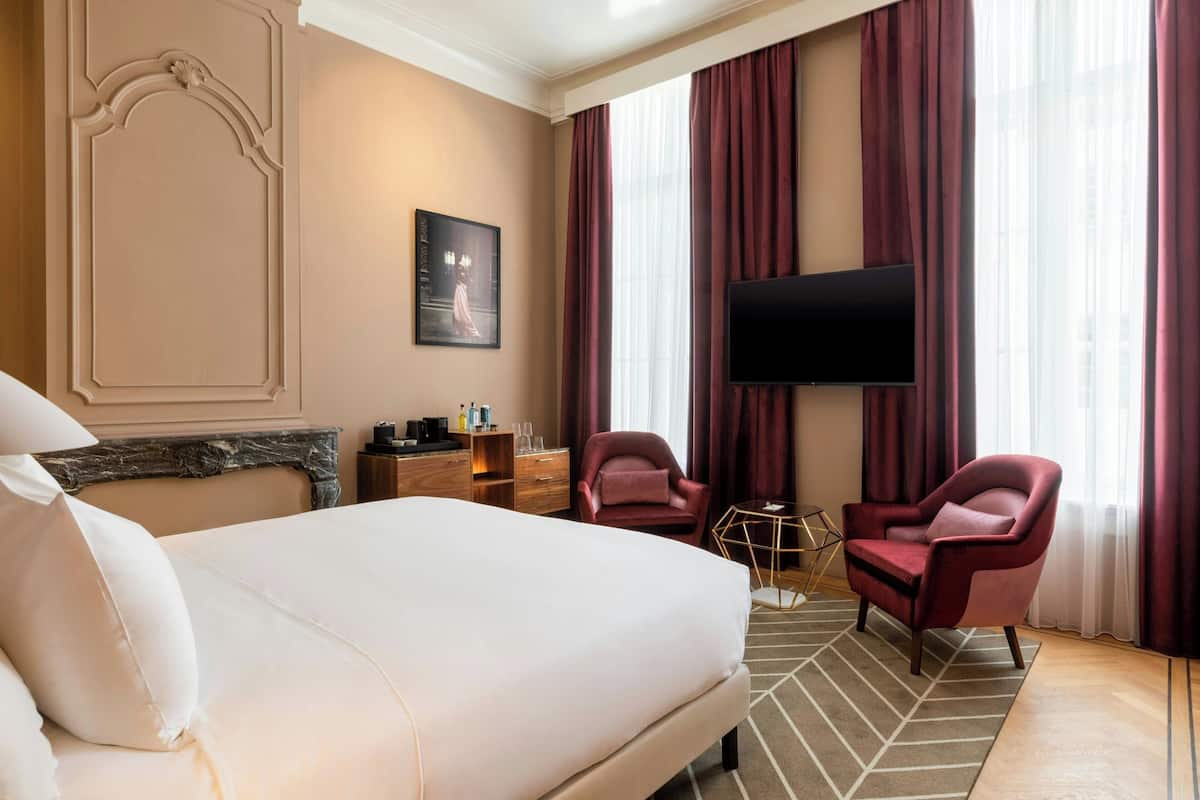 Ein Hotelzimmer mit einem großen Bett, zwei Sesseln, einem Flachbildfernseher, einem Marmor-Kamin und einem großen Fenster mit Vorhängen.