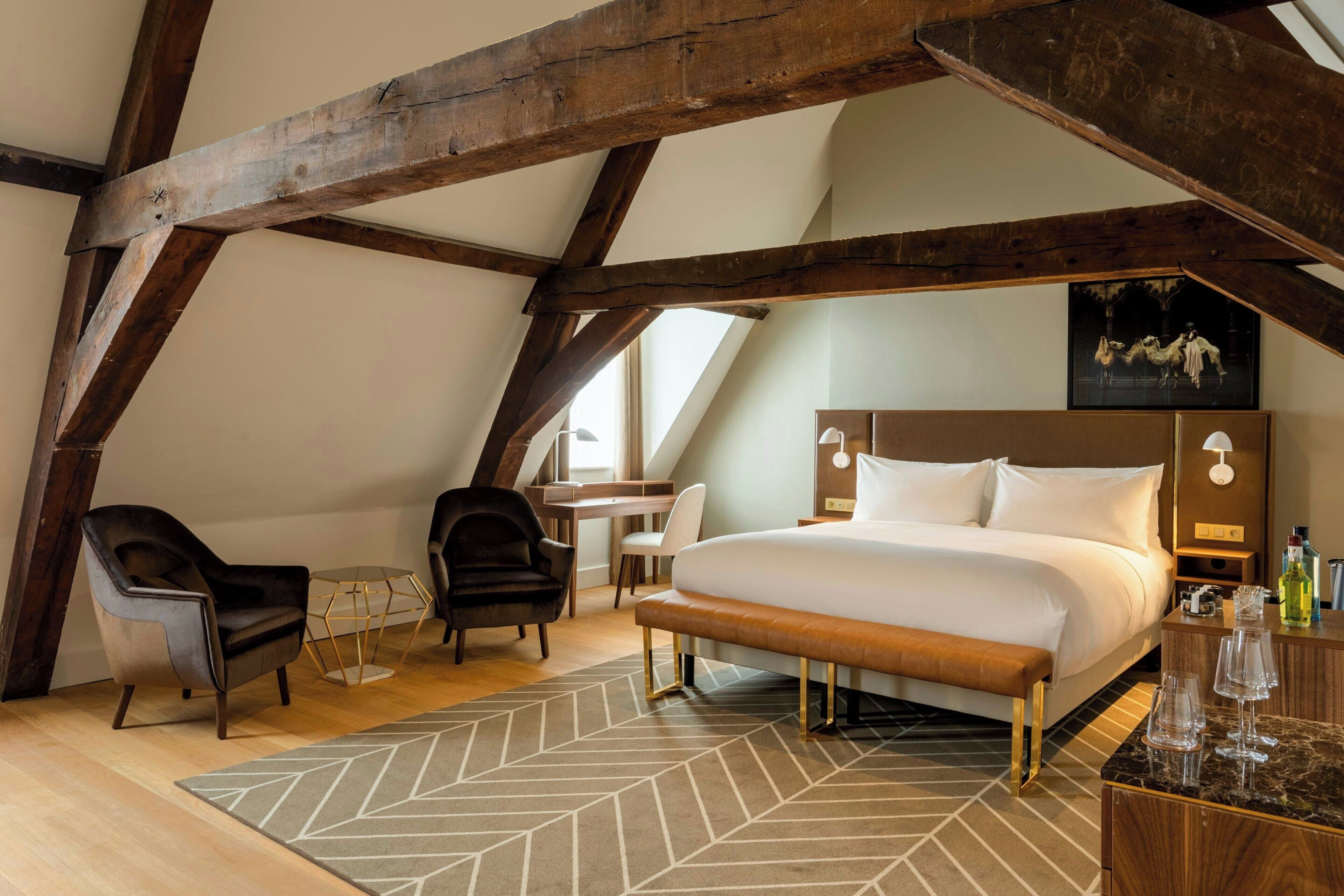 A bedroom with a large bed, wooden beams, a desk, two chairs, and a small table with a lamp.