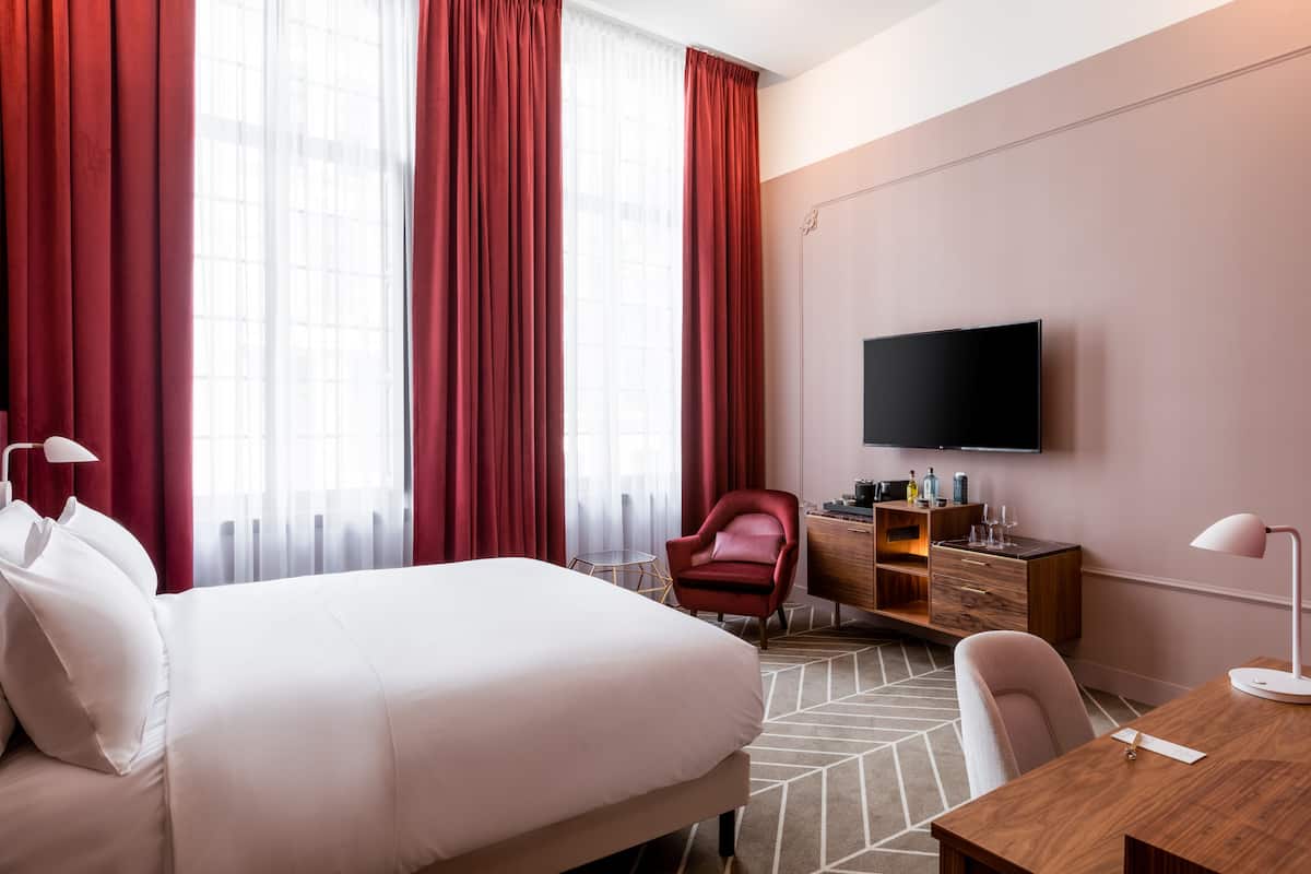 A hotel room with a large bed, a desk with a lamp, a TV mounted on the wall, and a red chair.