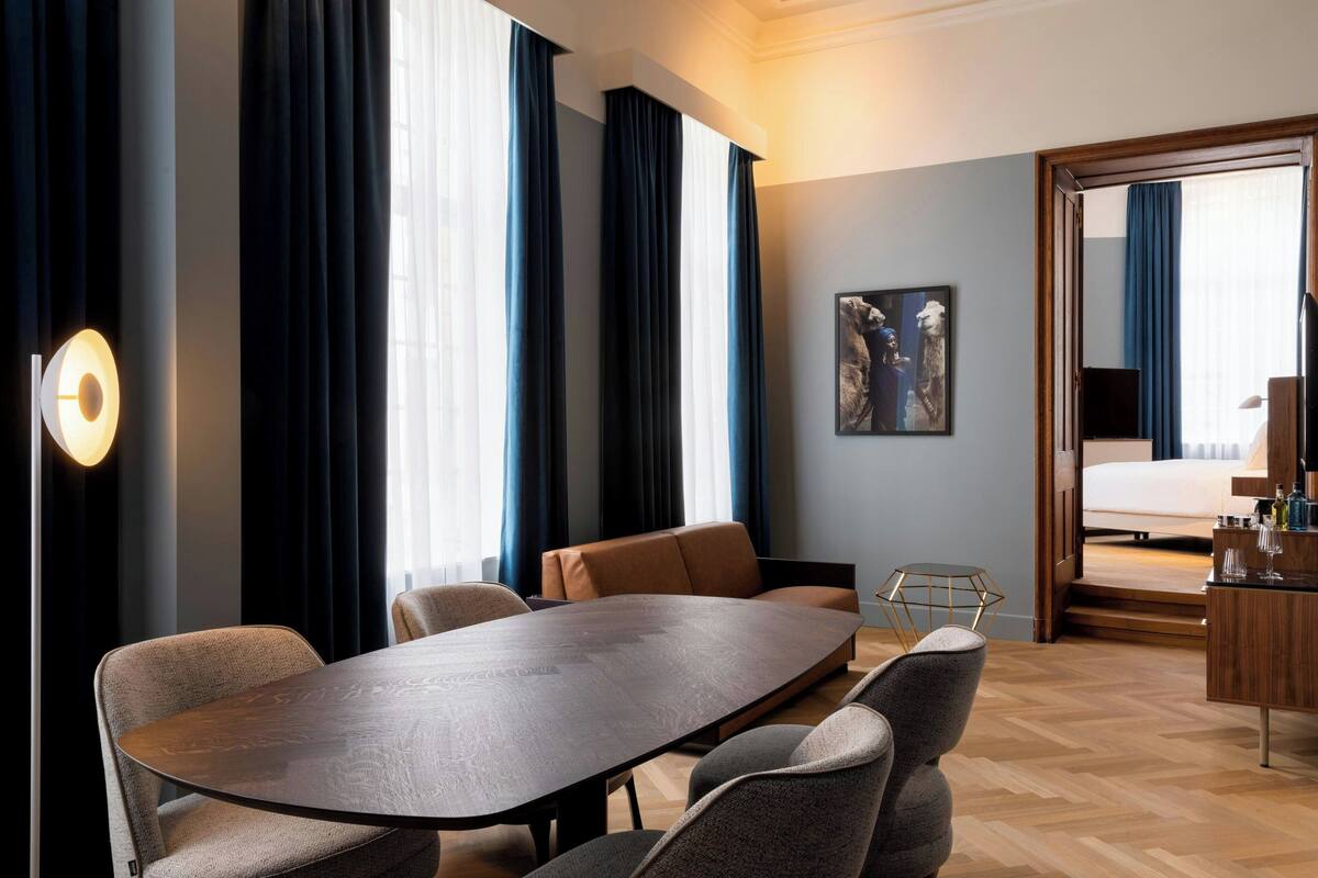 Ein modernes Hotelzimmer mit einem großen Holztisch, gemütlicher Sitzgelegenheit, einem Bett mit Holz Kopfteil, einem kleinen Schreibtisch mit Spiegel und einem großen Fenster mit Vorhängen.