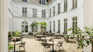 A courtyard with round tables and chairs, surrounded by white buildings with arched windows and a brick floor. - Sapphire House Antwerp, Autograph Collection (Antwerp)