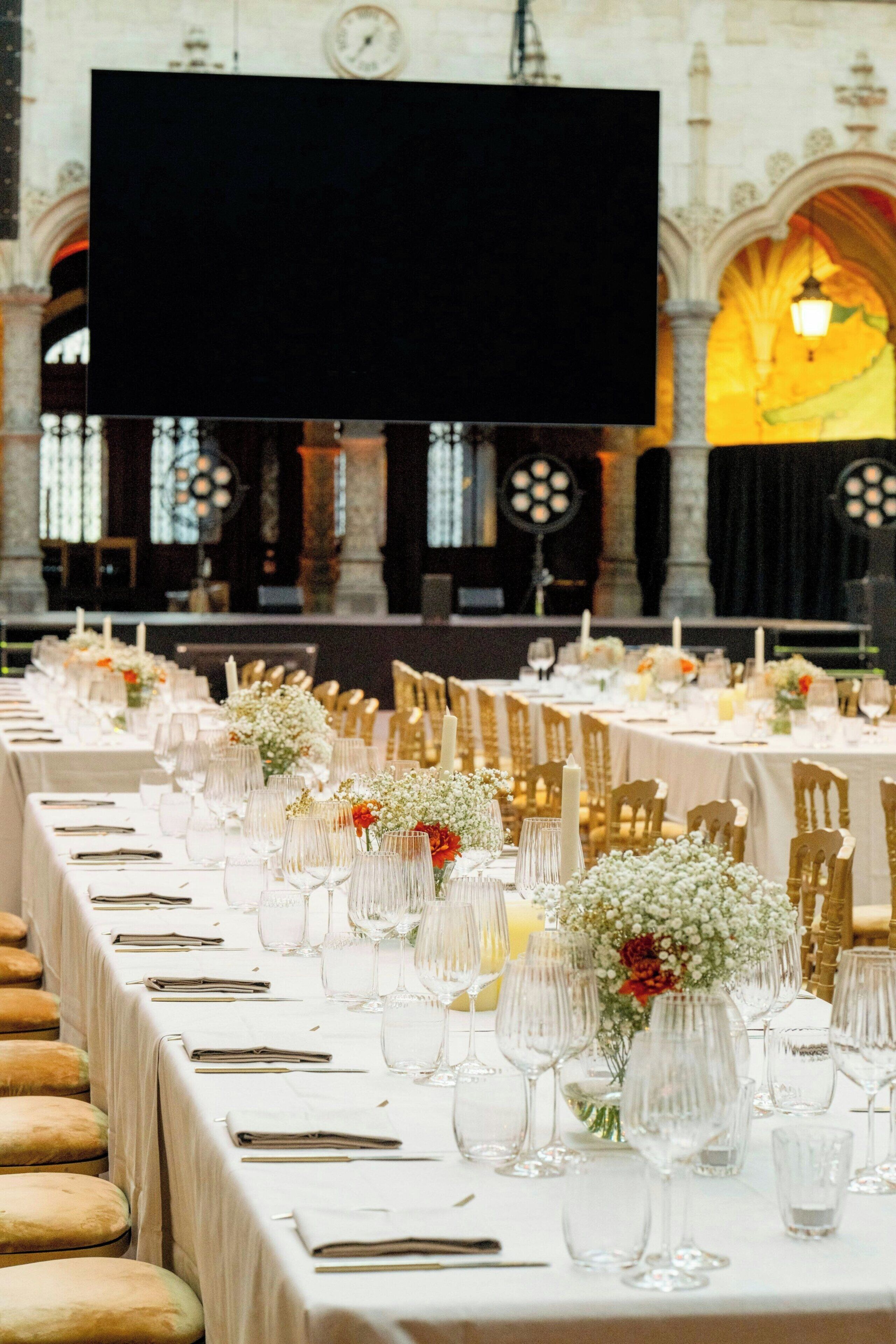 Un salón de banquetes con largas mesas dispuestas para un evento formal, con manteles blancos, sillas doradas y centros de mesa florales.