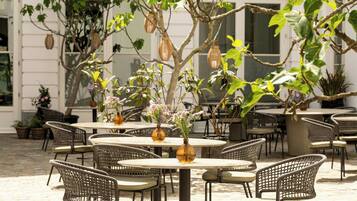 An outdoor patio with wicker chairs and tables, surrounded by potted plants and hanging lanterns.