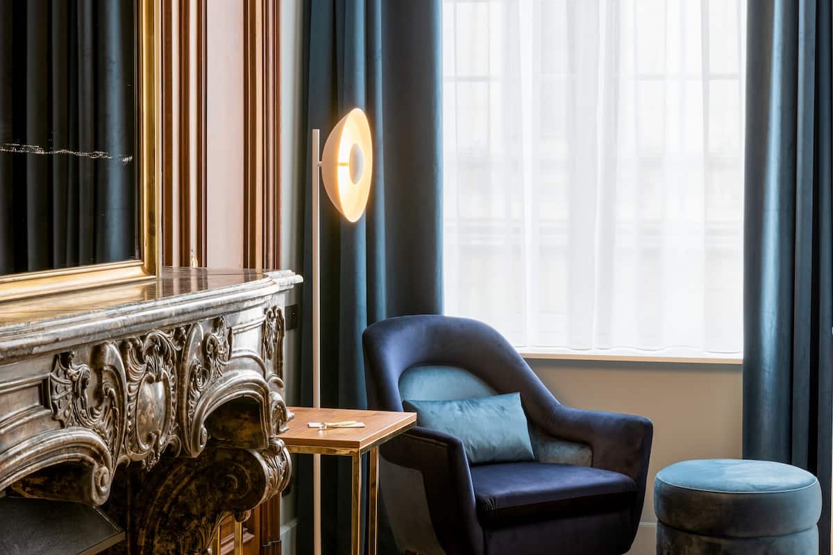 A room with a dark blue armchair, a matching ottoman, and a marble fireplace.