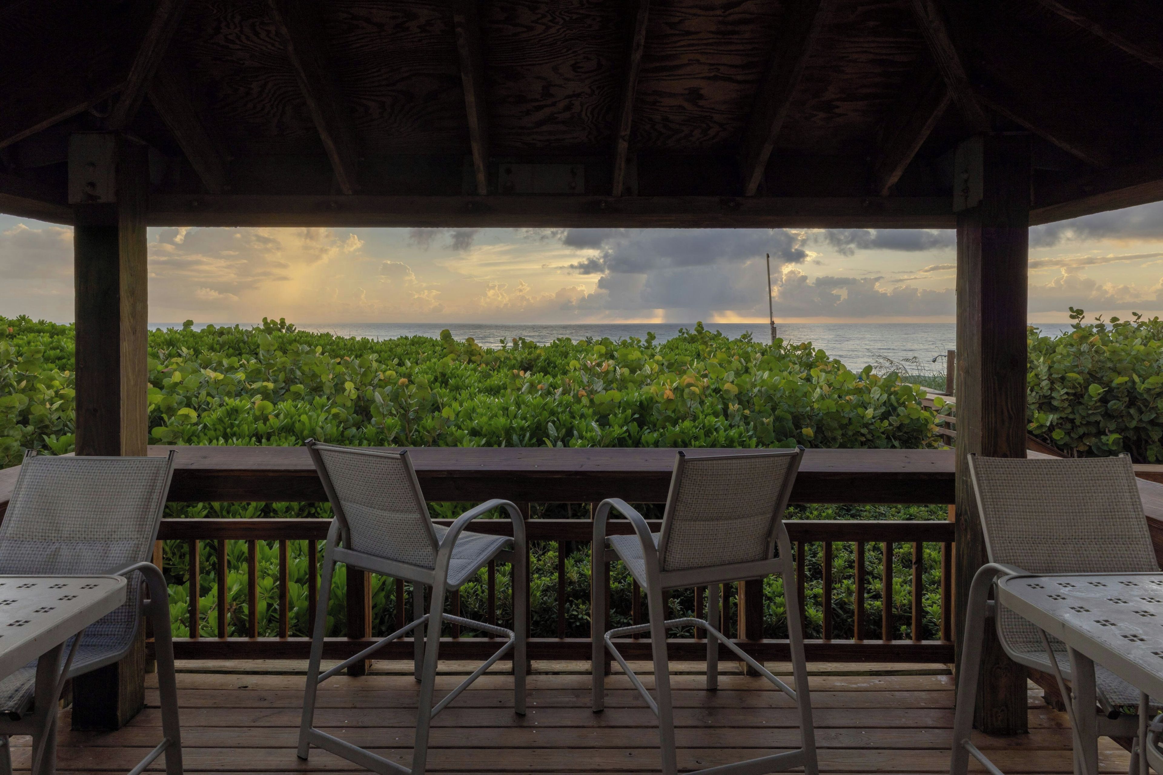 Un espacio de estar al aire libre techado con dos sillas y una mesa, con vistas a un paisaje verde exuberante y al mar.