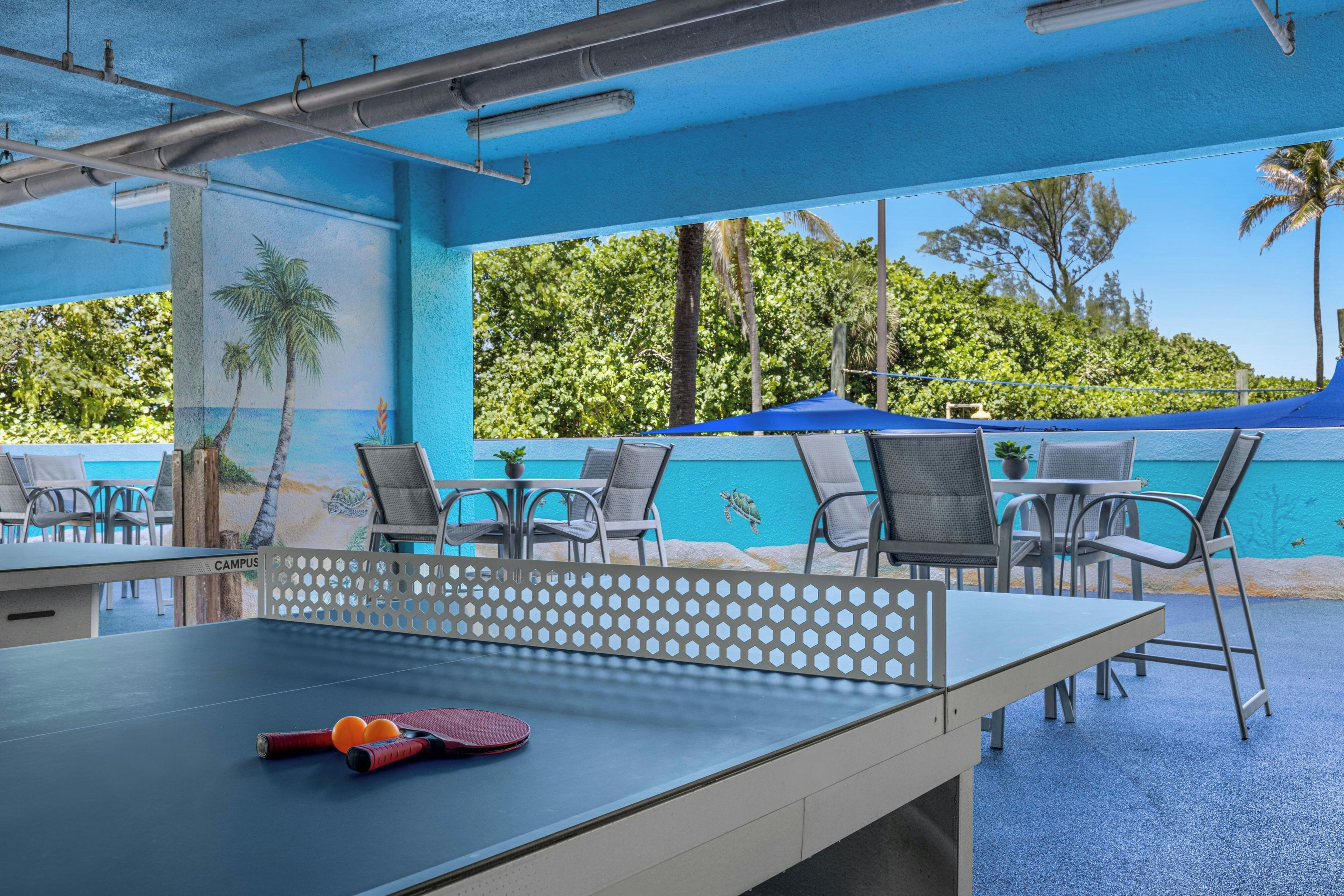Una mesa de ping pong al aire libre con red y paletas, rodeada de sillas, bajo un toldo azul con vistas a un paisaje tropical.