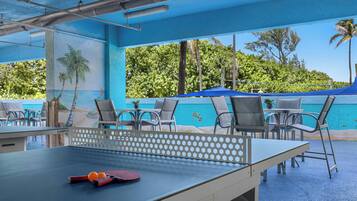 Una mesa de ping pong al aire libre con red y paletas, rodeada de sillas, bajo un toldo azul con vistas a un paisaje tropical.