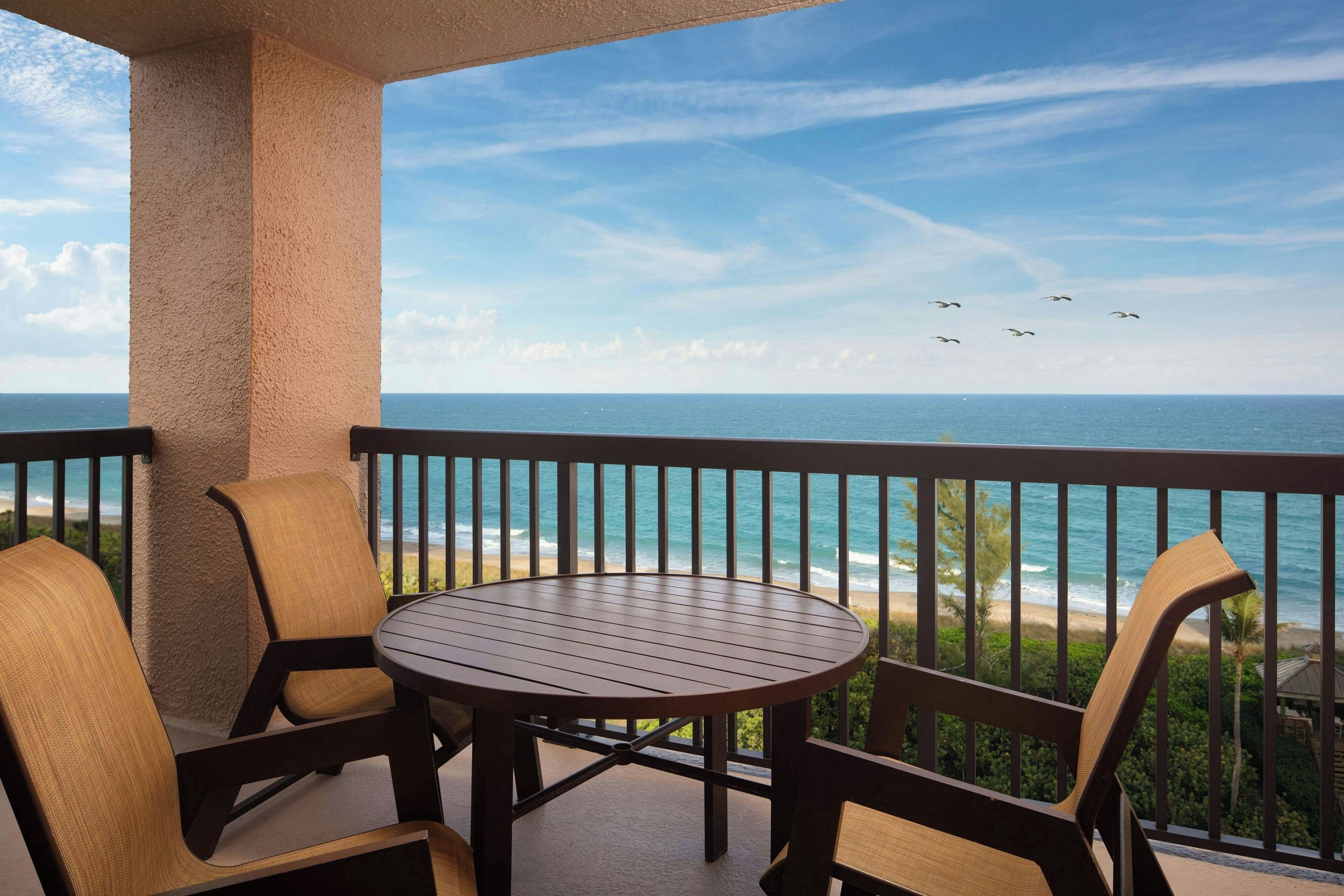 Un balcon avec une table et des chaises, donnant sur une plage où l’on peut admirer les vagues et un ciel dégagé.