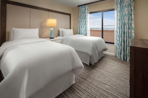 Quarto de hotel com duas camas, uma mesa de cabeceira com abajur, vista para o mar através de uma janela com cortinas estampadas e piso coberto por carpete.