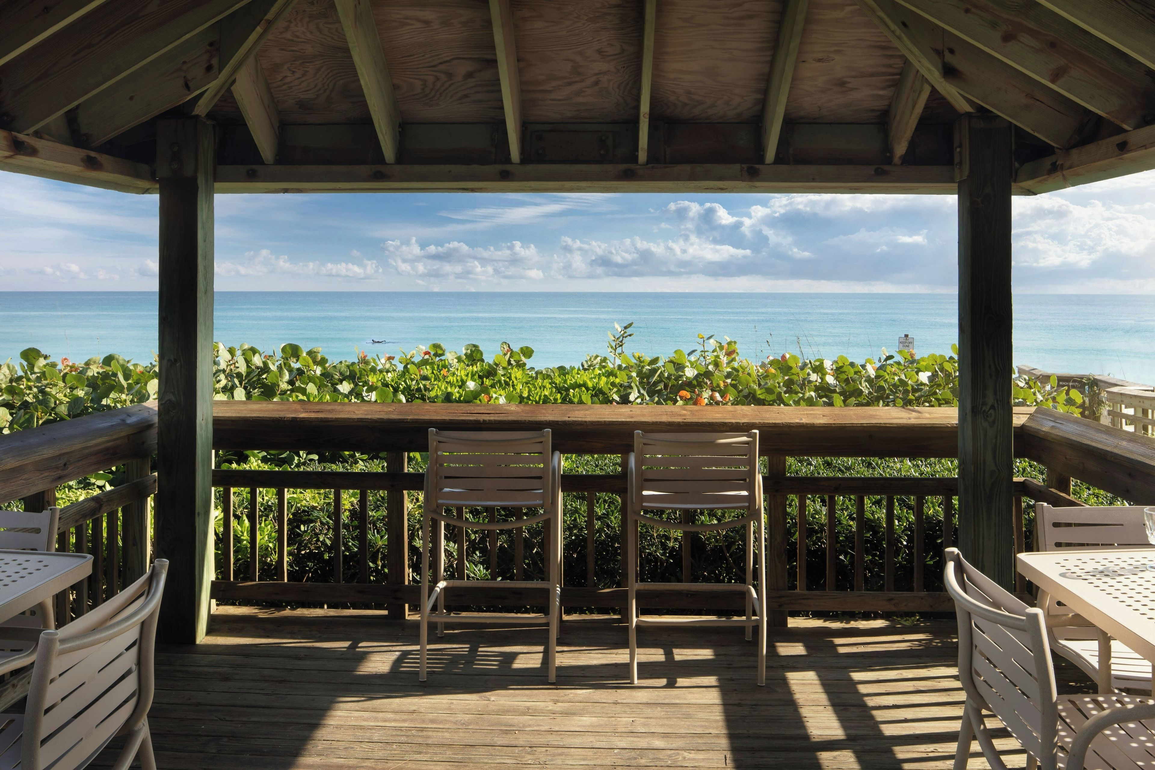 Una terraza de madera con dos sillas y una mesa, con vista a una playa y agua azul cristalina, y un letrero que indica zona de playa.