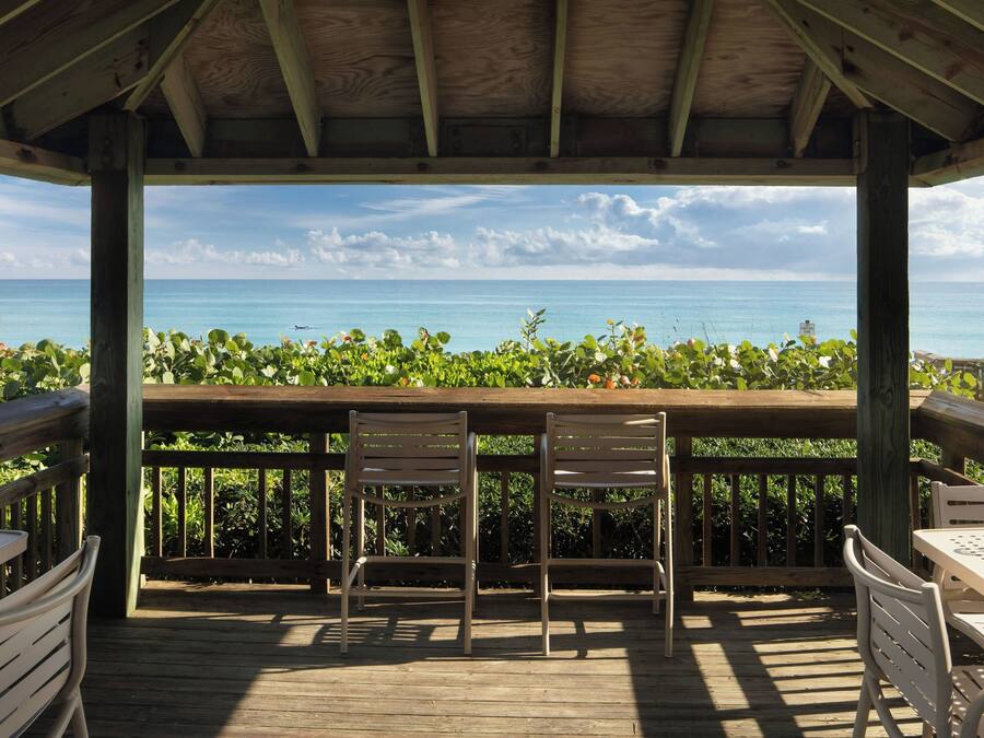A wooden deck with two chairs and a table, overlooking a beach with clear blue water and a sign indicating a beach area.