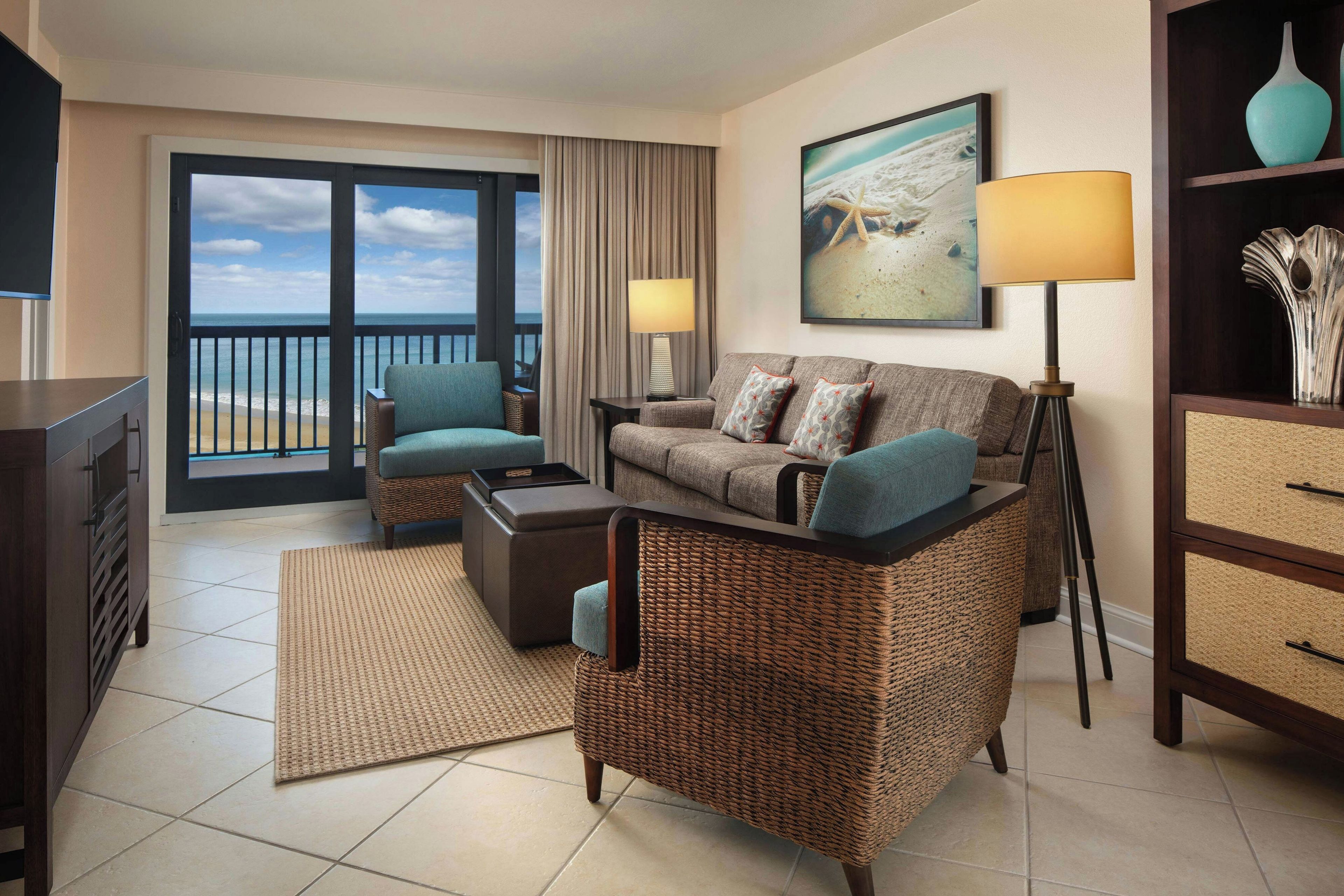 A hotel room with a balcony, a sofa set, a coffee table, a side table, a lamp, and a painting of a beach scene.
