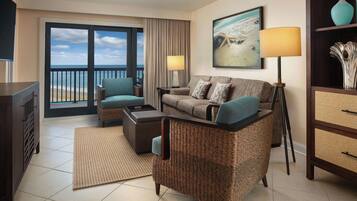 A hotel room with a balcony, a sofa set, a coffee table, a side table, a lamp, and a painting of a beach scene.