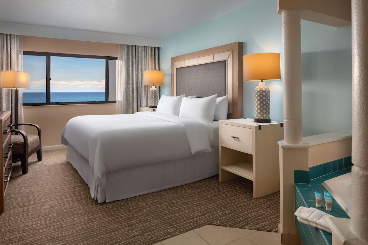 A hotel room with a large bed, two bedside tables, a desk, and a view of the ocean.