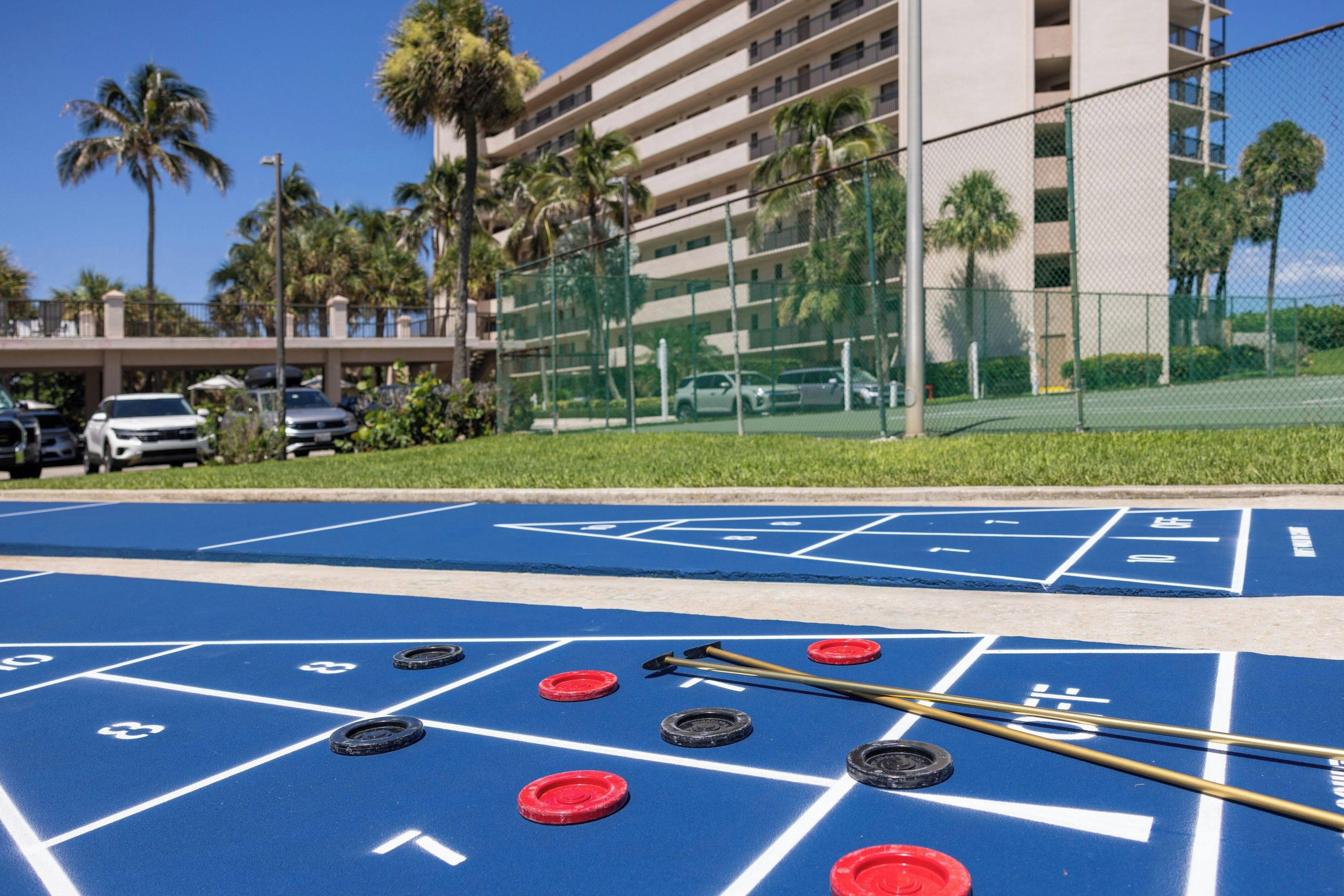 A shuffleboard court with pucks and sticks, a parking lot with cars, and a building with balconies and palm trees in the background.