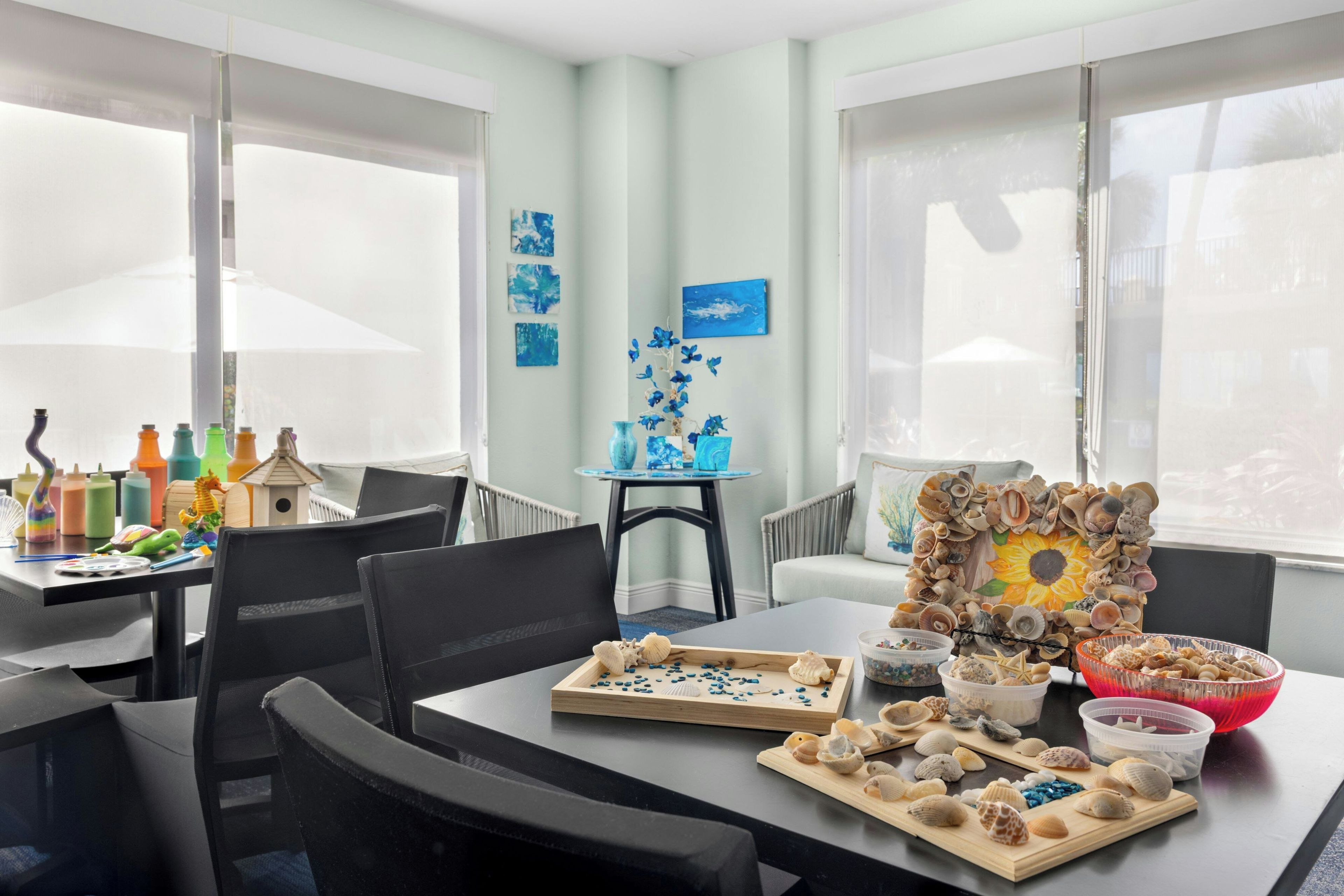 Zona pranzo con tavolo apparecchiato per due, con un centrotavola di conchiglie e un quadro di girasole.