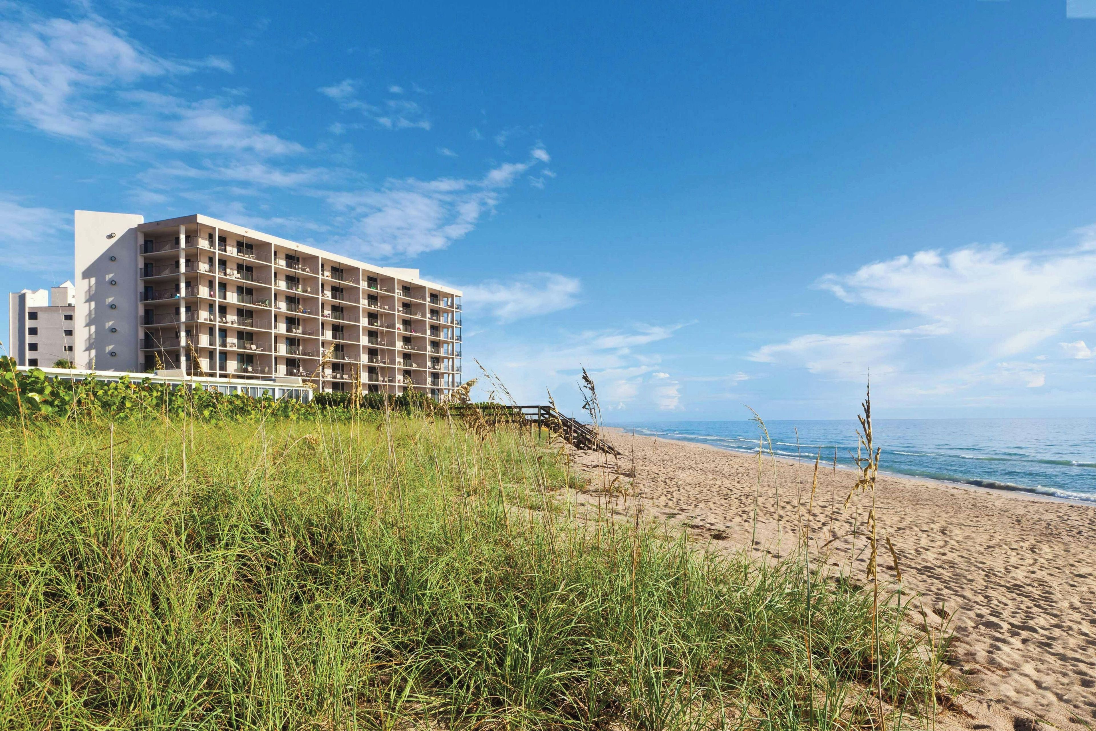 A beachfront hotel with multiple floors and balconies, situated on a sandy beach with grass in the foreground.