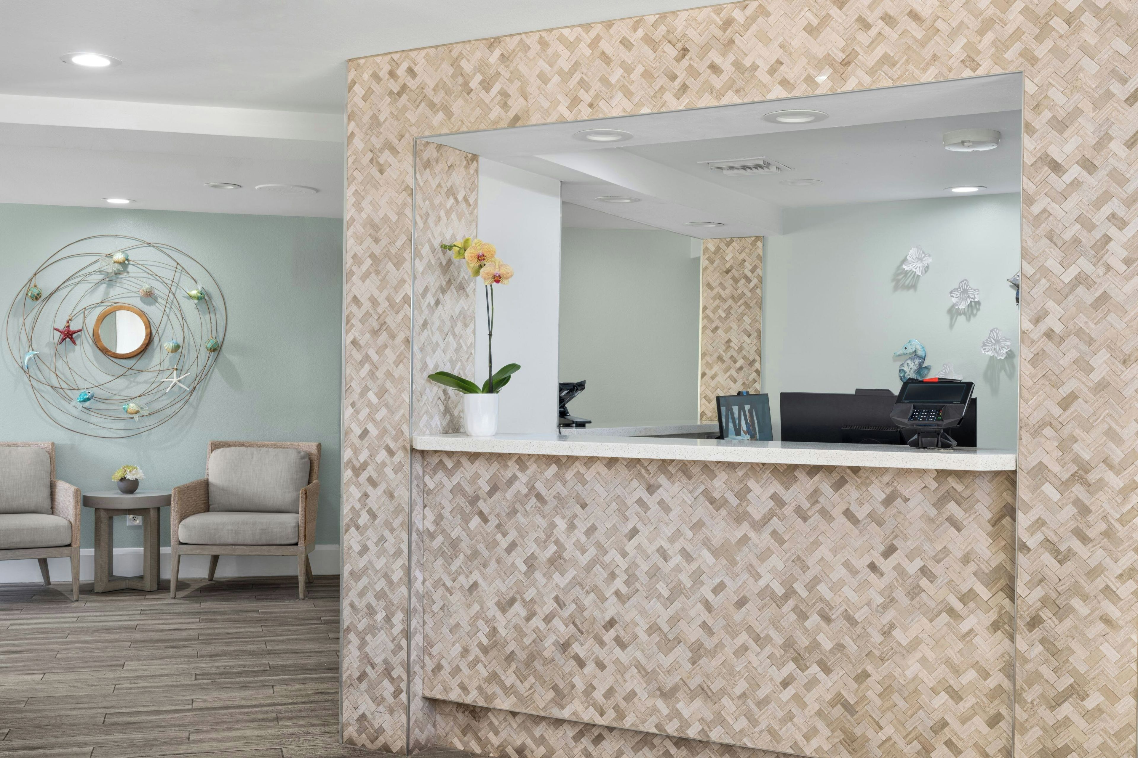 A reception area with a herringbone patterned wall, a reception desk, two chairs, a small table with a vase, and a decorative wall piece.