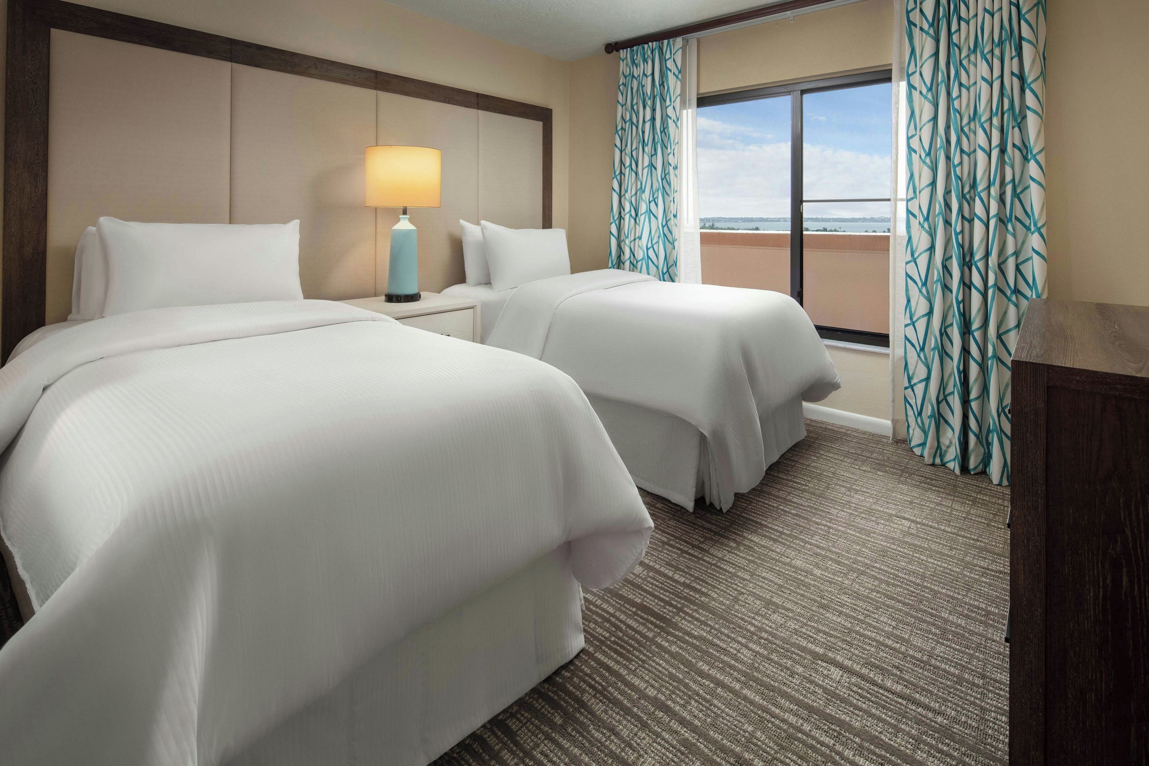 A hotel room with two beds, a nightstand with a lamp, and a view of the sea through the window.