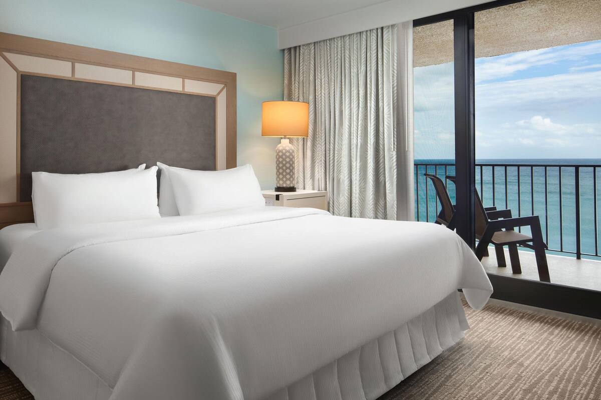 A hotel room with a large bed, a nightstand with a lamp, a chair, and a view of the ocean.