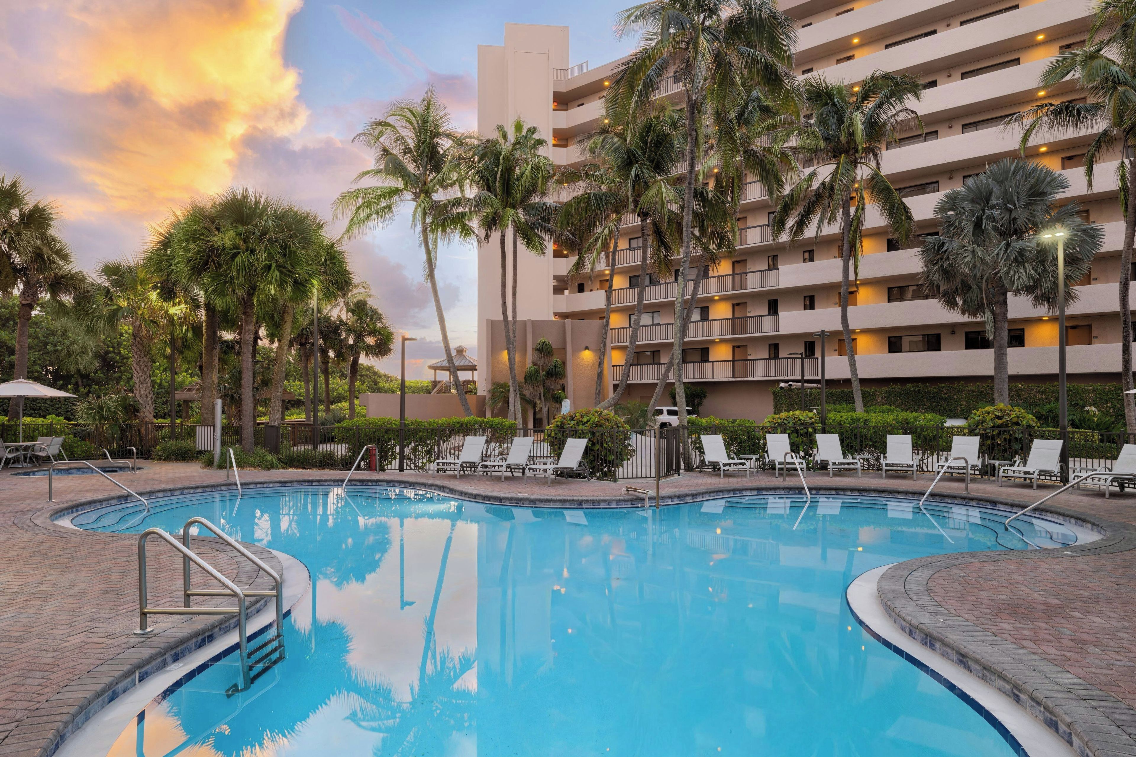 A hotel pool area with a clear sky, palm trees, and a multi-story building in the background.