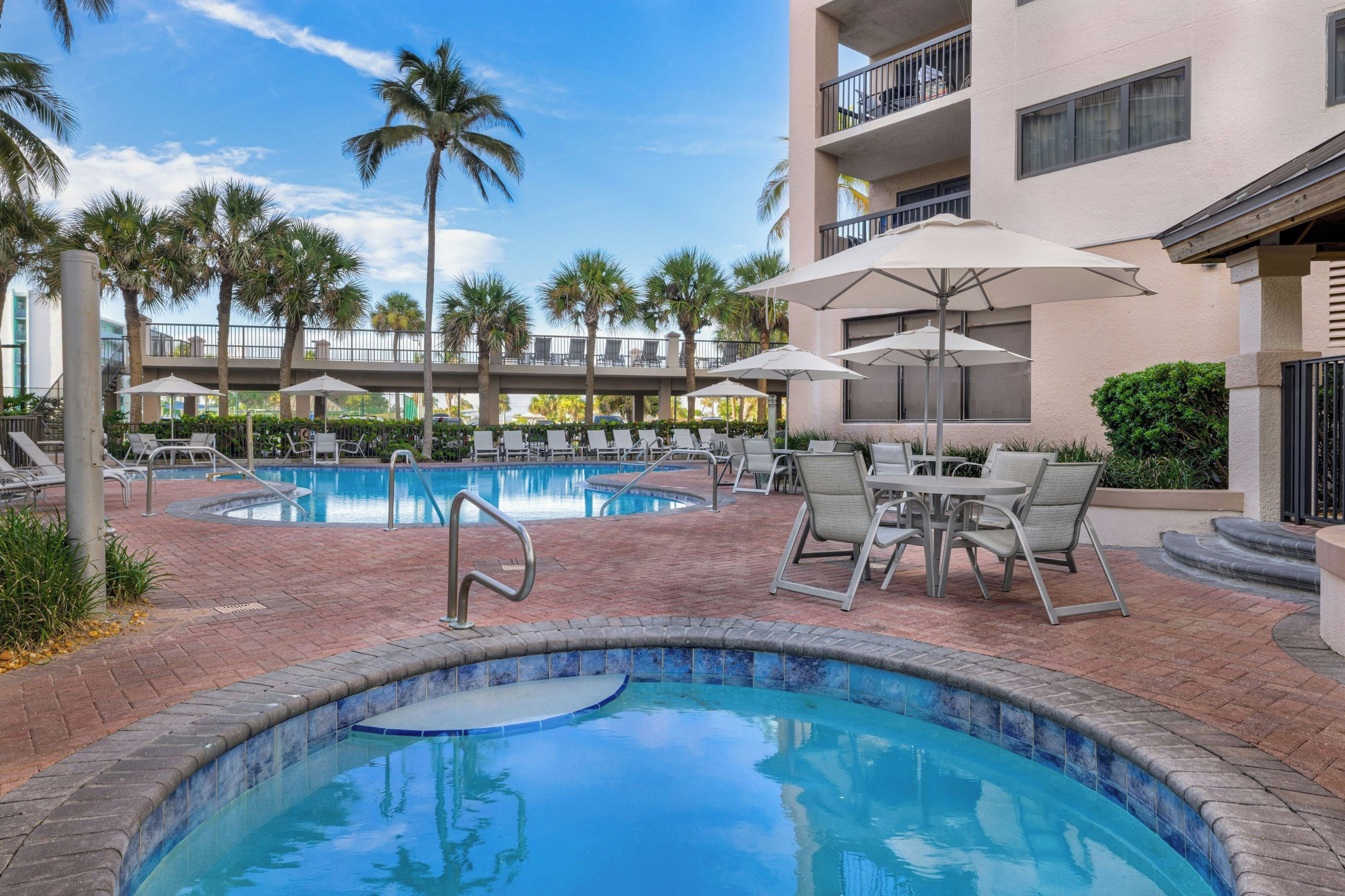 A pool area with lounge chairs and umbrellas, surrounded by palm trees and a building with balconies.