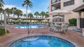 A pool area with lounge chairs and umbrellas, surrounded by palm trees and a building with balconies.