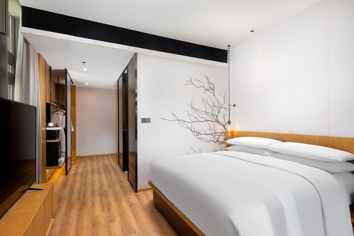 A modern hotel room with a wooden floor, a large bed with white bedding, a wardrobe, and a wall-mounted artwork.