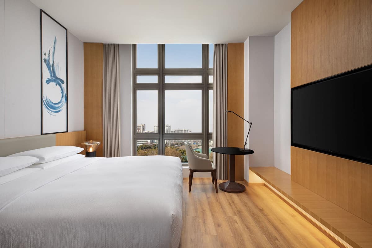 A modern hotel room with a large bed, a TV, a desk, and a view of the city through large windows.